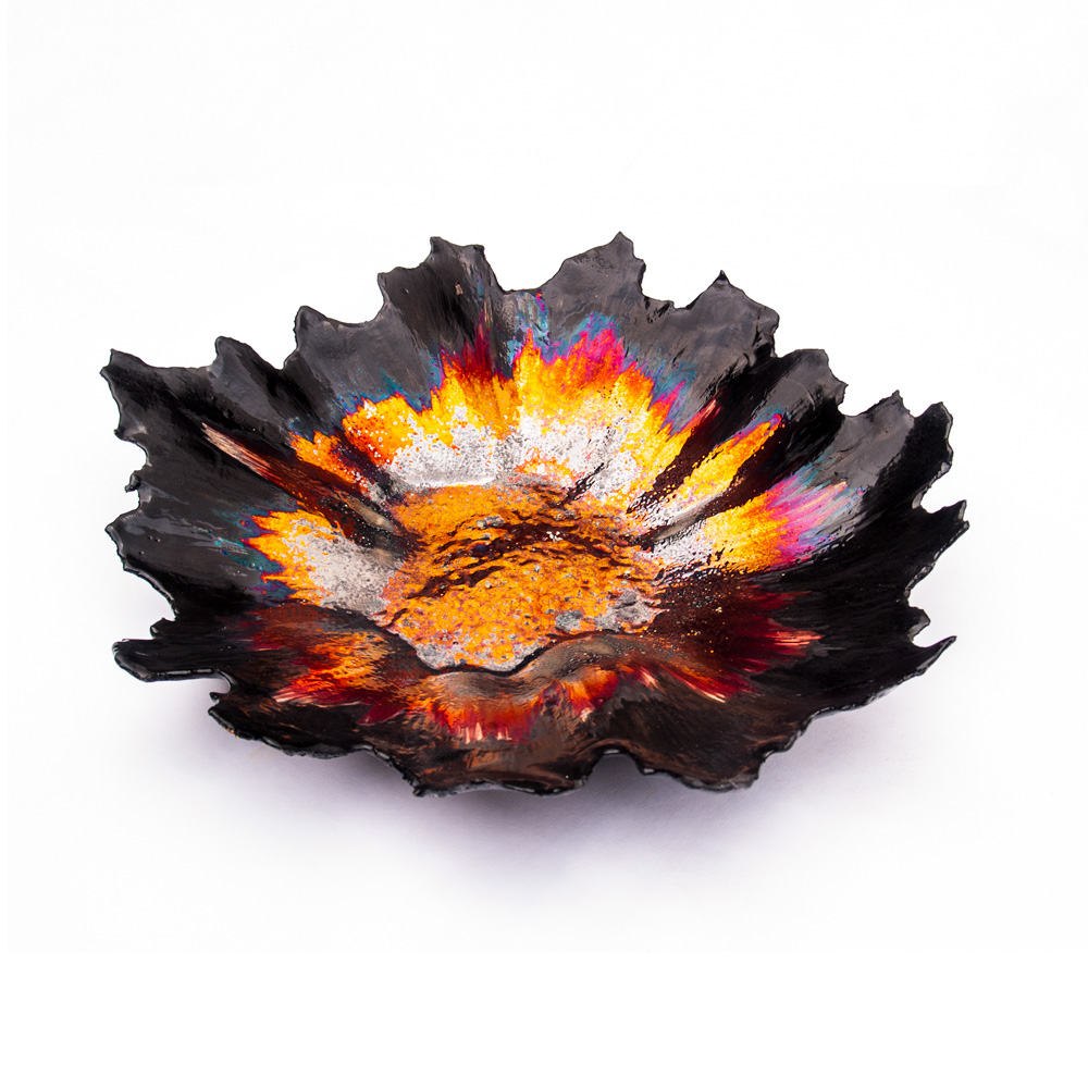 A three-quarter angle view of a handcrafted dark ceramic vessel from the Volcano series by Alireza Rostami, featuring jagged edges and a glowing metallic luster glaze in orange, gold, and magenta on a plain white background.