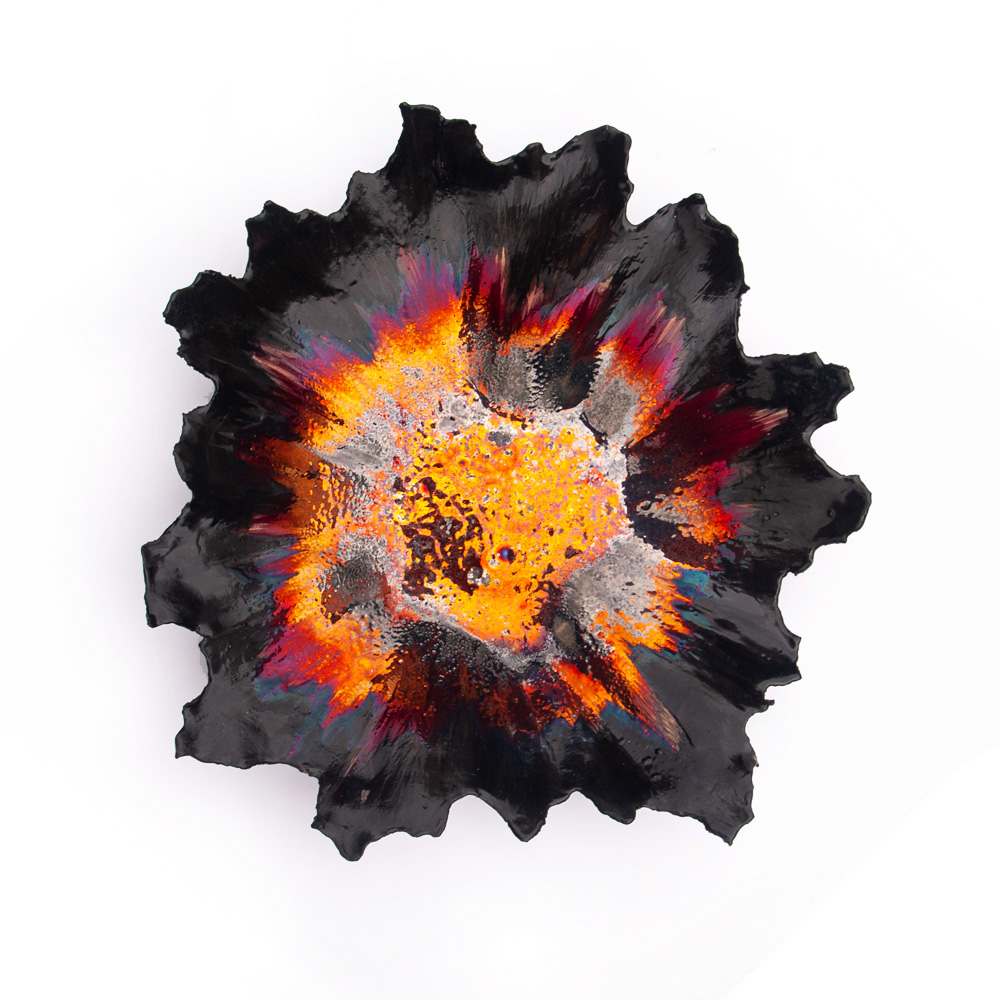 A high-angle top-down view of a handcrafted black ceramic vessel from the Volcano collection by Alireza Rostami, displaying a radiating burst of fiery orange, magenta, and yellow metallic luster glazes at its center on a white background.