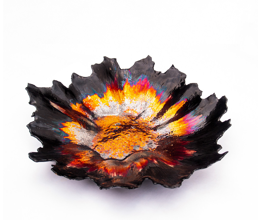 A three-quarter angle view of a handcrafted dark ceramic vessel from the Volcano series by Alireza Rostami, featuring jagged edges and a glowing metallic luster glaze in orange, gold, and magenta on a plain white background.